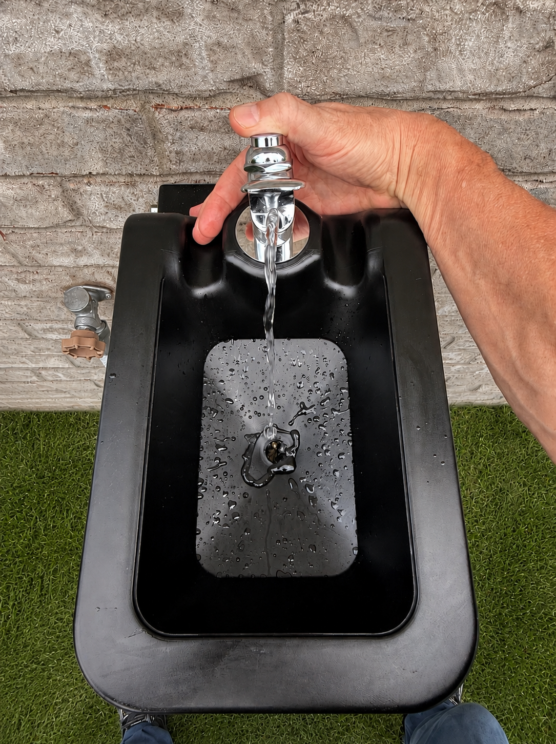 A black outdoor water fountain basin with a chrome faucet, shown with a person's hand turning on the water, set against a gray brick wall and green grass.