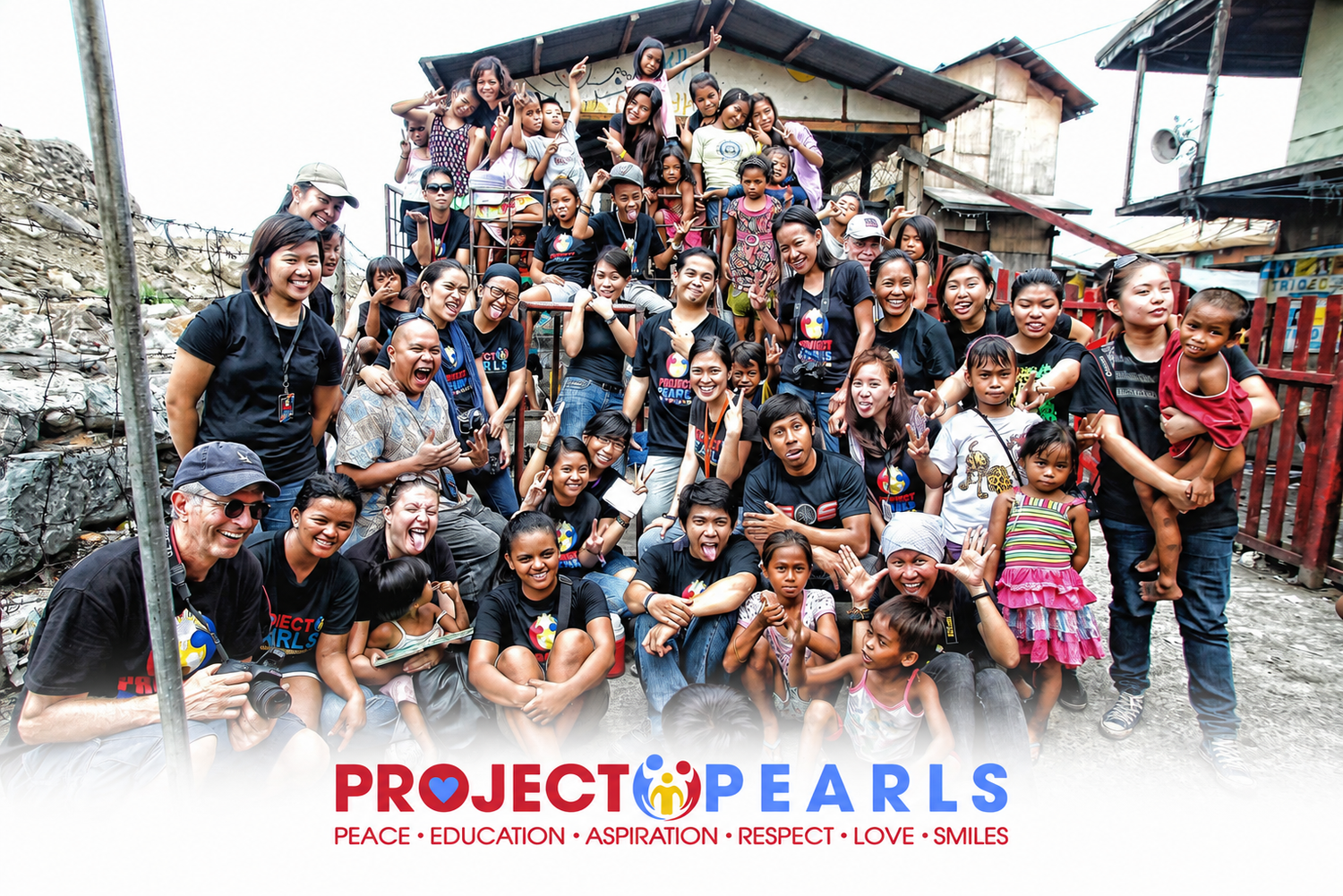 A large group of people posing together outdoors in front of a building, with a Project Pearls logo banner overlaid at the bottom. The scene includes adults, children, and a woman holding a baby.