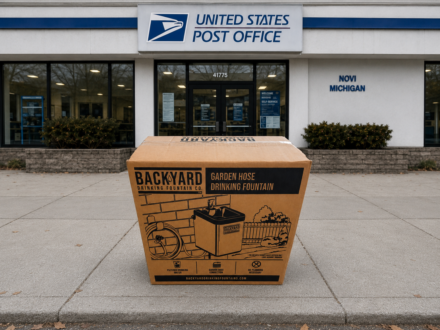 A large cardboard shipping box for a backyard garden hose drinking fountain sits on a concrete sidewalk in front of a United States Post Office building in Novi Michigan.